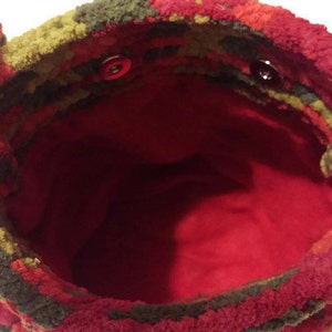 Puede incluir: Una bolsa de crochet roja, verde y marr&oacute;n con un forro rojo y dos botones rojos. La bolsa est&aacute; hecha de un material suave y peludo.