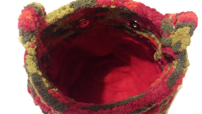 Puede incluir: Una bolsa de crochet roja, verde y marr&oacute;n con un forro rojo y dos botones rojos. La bolsa est&aacute; hecha de un material suave y peludo.