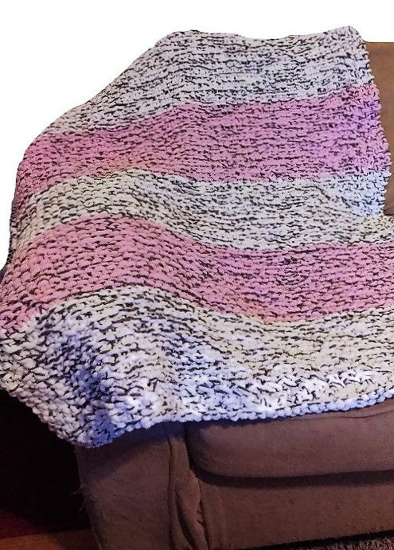 May include: A soft, chunky knit throw blanket with stripes of white, pink, and gray. The blanket is draped over a brown couch.
