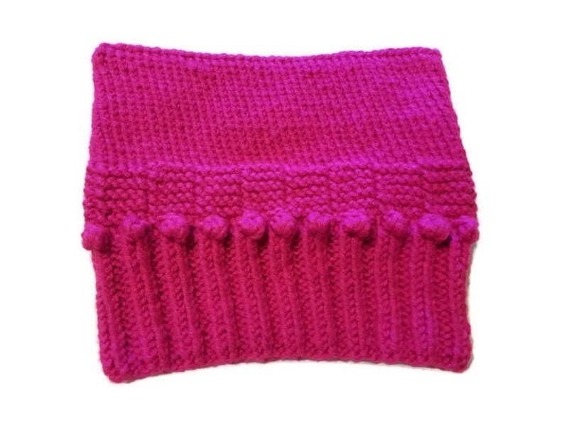 May include: A vibrant fuchsia-colored knitted cowl. The cowl features a ribbed bottom section with small bobbles, a textured middle section, and a smooth top. This handmade accessory is perfect for staying warm in cold weather.