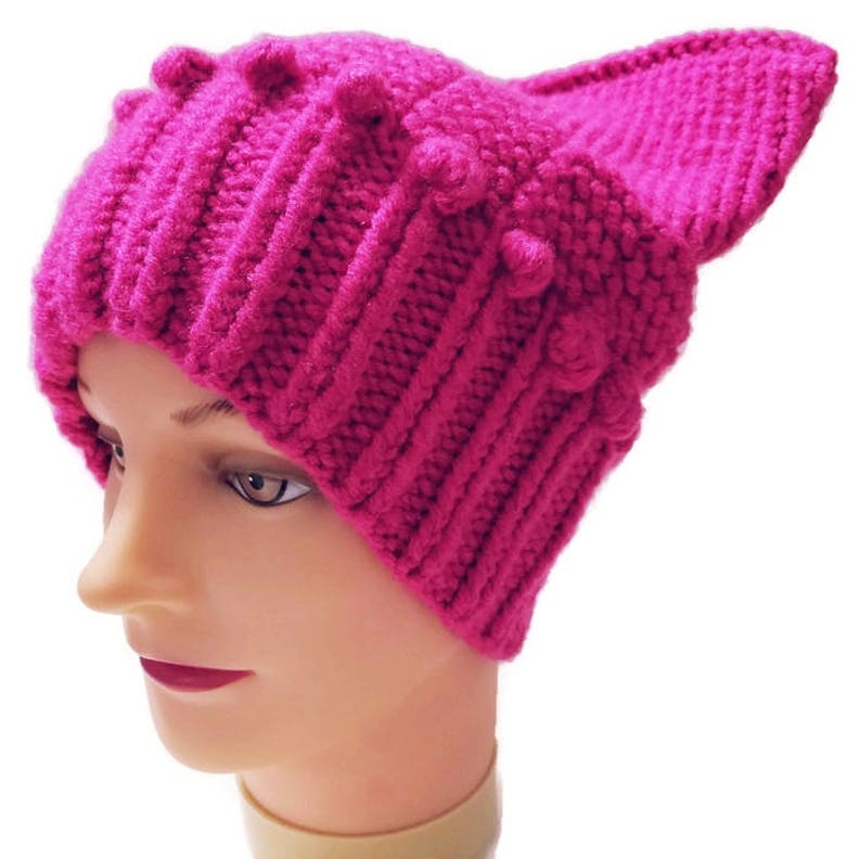 May include: A bright pink knitted hat with ear-like protrusions. The hat features a ribbed design and small, decorative bobbles. The hat is displayed on a mannequin head, showcasing its shape and texture. The hat is a vibrant accessory.