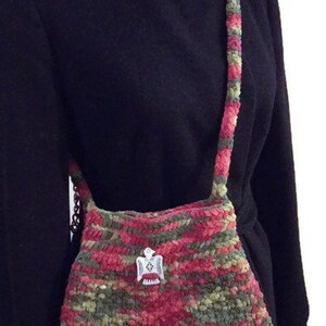 Puede incluir: Un bolso de hombro de ganchillo en tonos rosa, verde y marr&oacute;n. El bolso tiene un dije de p&aacute;jaro plateado.