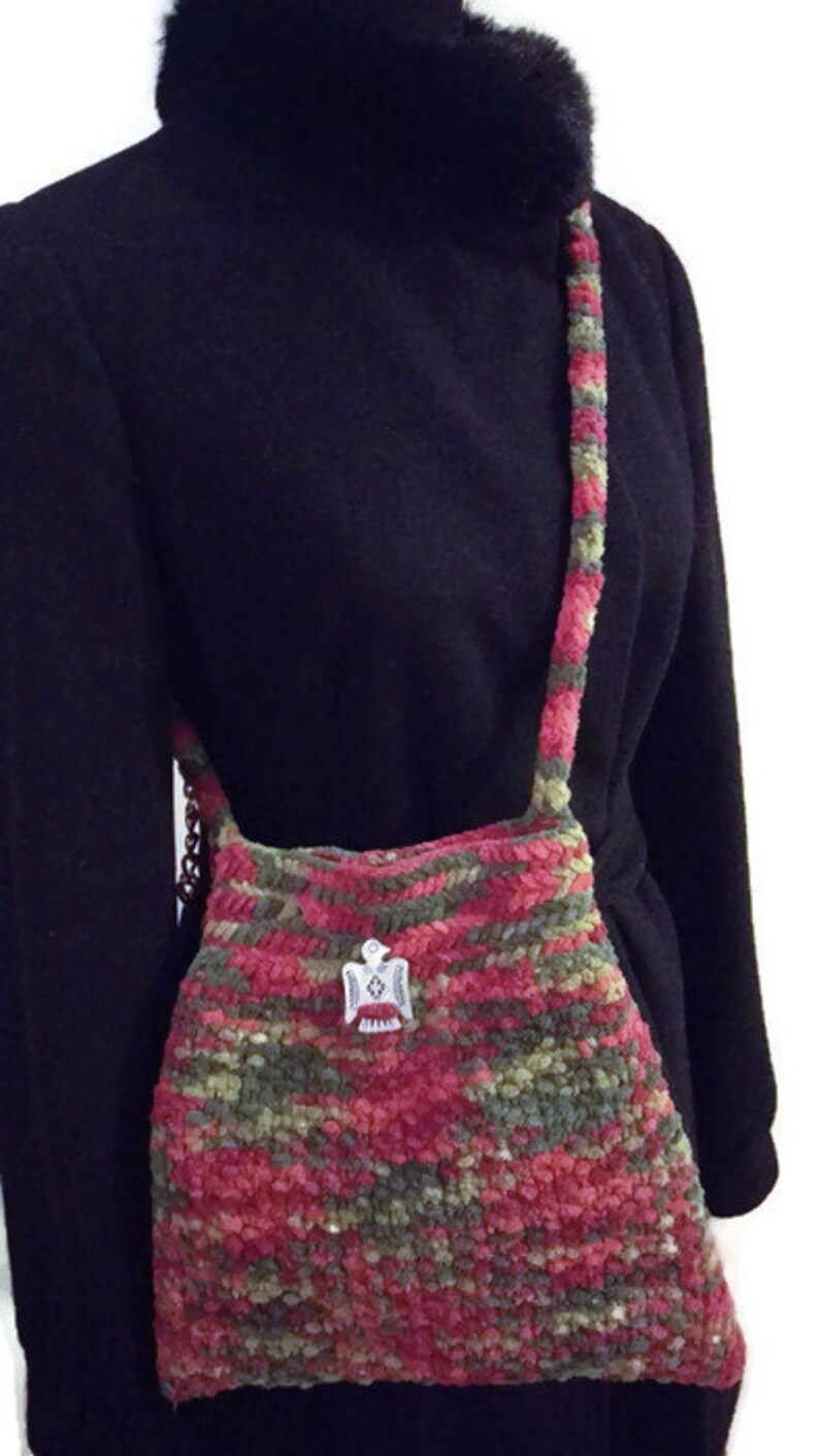 Puede incluir: Un bolso de hombro de ganchillo en tonos rosa, verde y marr&oacute;n. El bolso tiene un dije de p&aacute;jaro plateado.