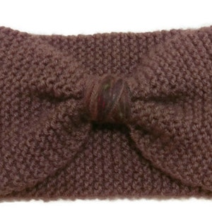 K&ouml;nnte beinhalten: Ein gestricktes Stirnband mit einer gro&szlig;en Schleife in der Mitte. Das Stirnband ist dunkel-mauvefarben und die Schleife ist in einem dunkleren Mauve-Ton mit einem kleinen mehrfarbigen Muster.