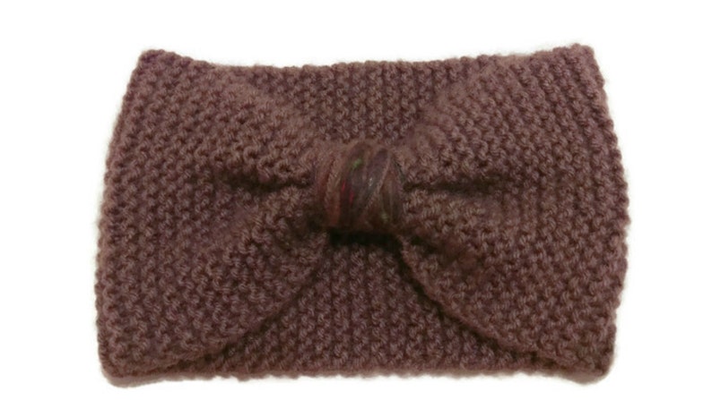 K&ouml;nnte beinhalten: Ein gestricktes Stirnband mit einer gro&szlig;en Schleife in der Mitte. Das Stirnband ist dunkel-mauvefarben und die Schleife ist in einem dunkleren Mauve-Ton mit einem kleinen mehrfarbigen Muster.