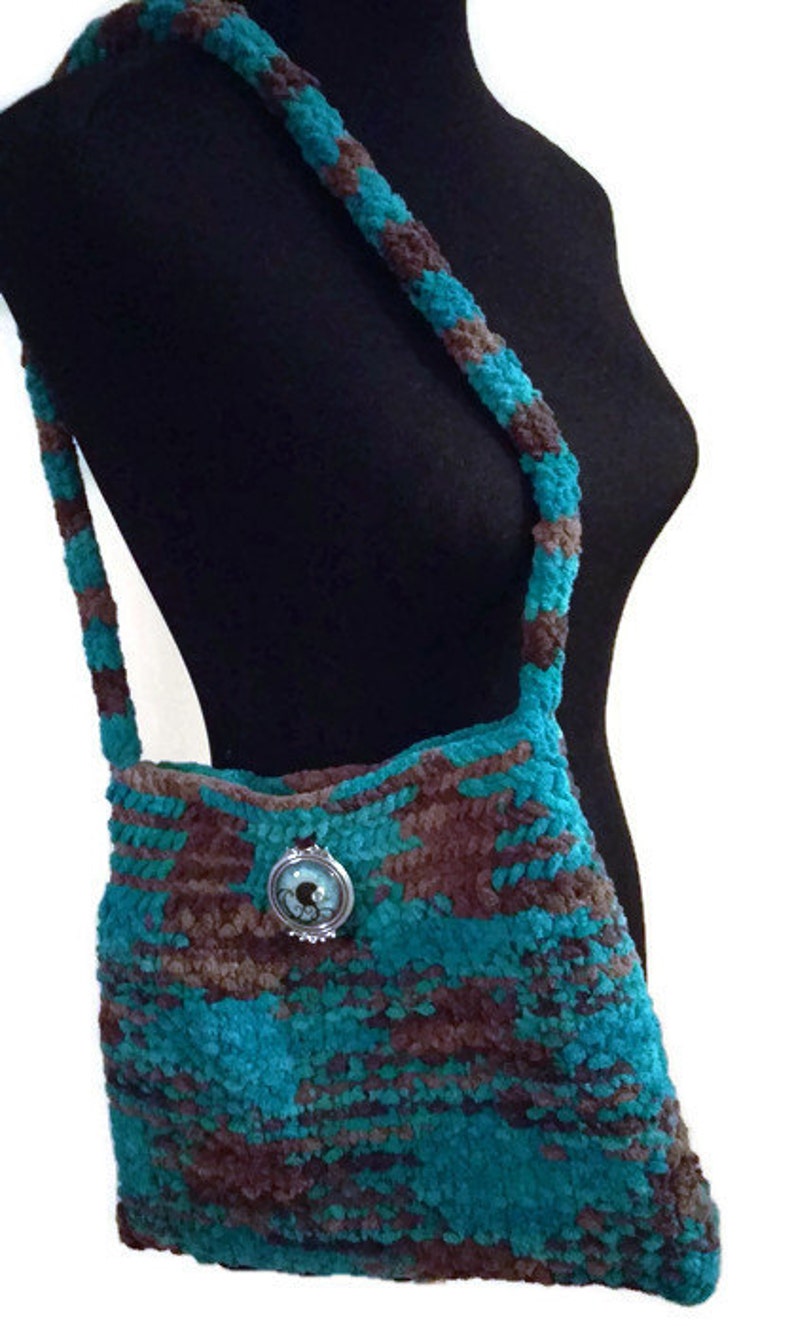 May include: A teal and brown crocheted shoulder bag with a decorative metal button. The bag has a long strap and is made of a soft, fuzzy yarn.