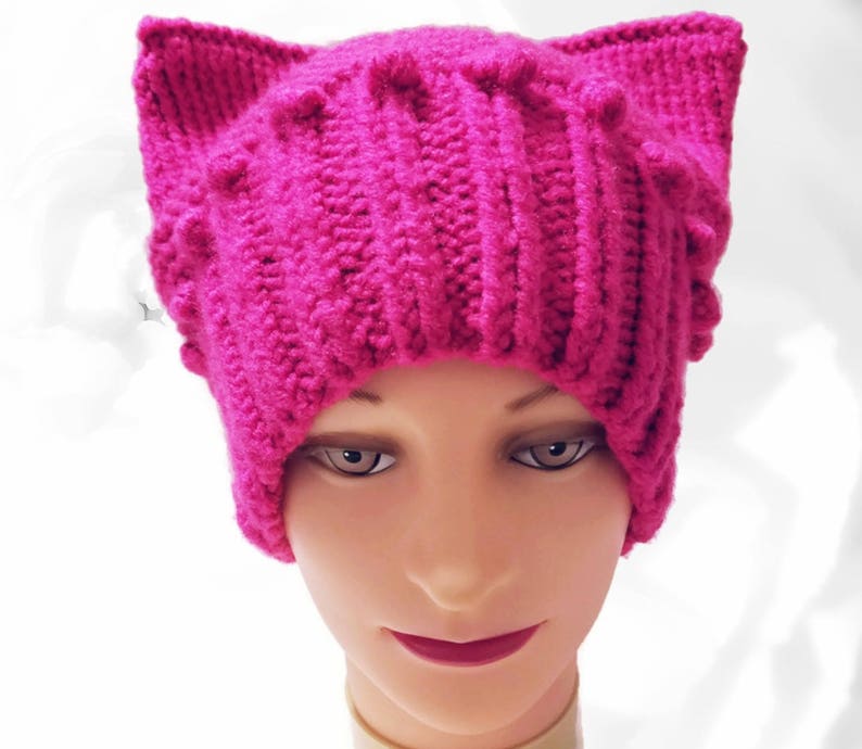 May include: A vibrant fuchsia-colored knitted hat with cat ears. The hat features a ribbed design and small, textured bobbles. The ears stand upright, adding a playful touch to the headwear. This handmade hat is a cozy and stylish accessory.