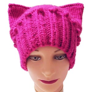 May include: A vibrant fuchsia-colored knitted hat with cat ears. The hat is made of thick yarn, featuring a ribbed design on the main body and textured details on the ears. The hat is displayed on a mannequin head against a white background.