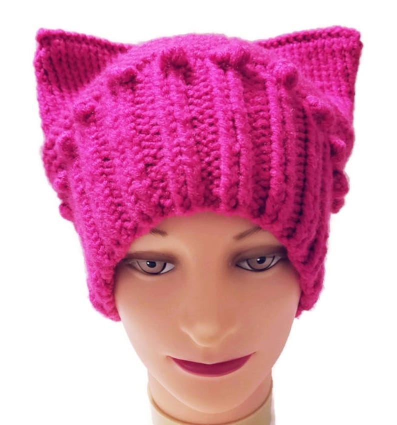 May include: A vibrant fuchsia-colored knitted hat with cat ears. The hat is made of thick yarn, featuring a ribbed design on the main body and textured details on the ears. The hat is displayed on a mannequin head against a white background.