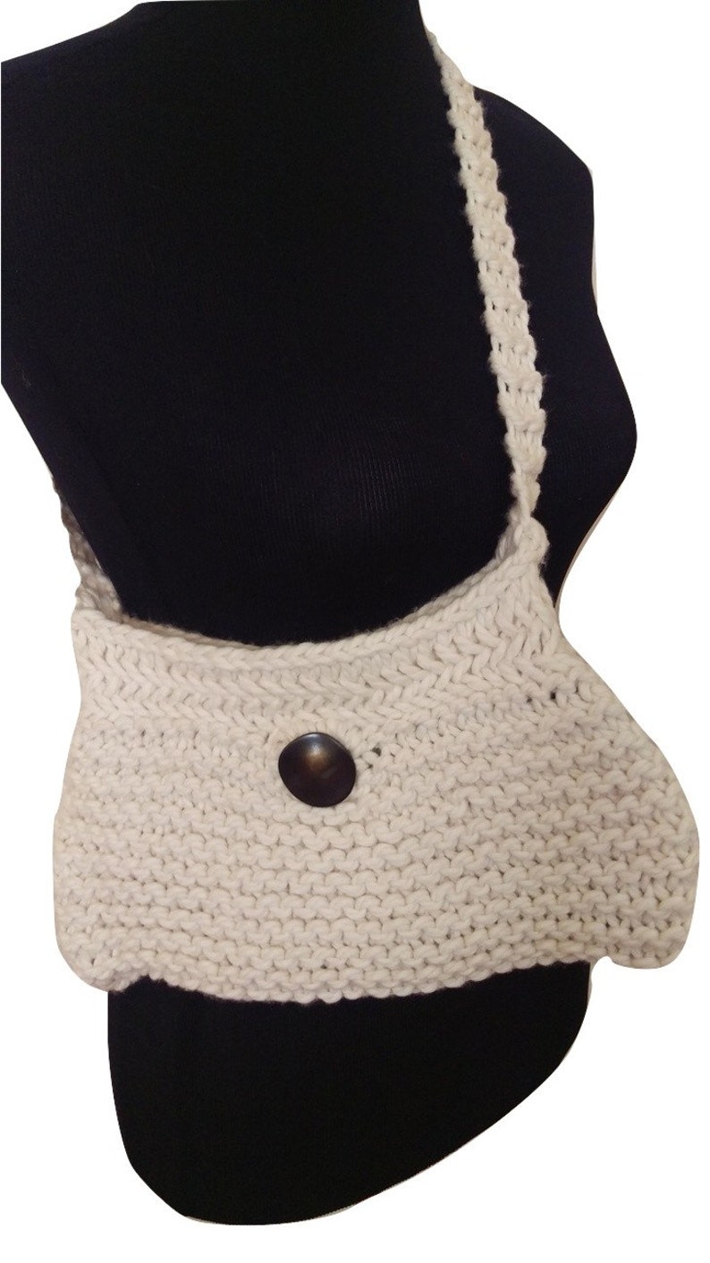 K&ouml;nnte beinhalten: Eine wei&szlig;e gestrickte Umh&auml;ngetasche mit braunem Knopfverschluss. Die Tasche ist aus dickem, strukturiertem Garn gefertigt und hat einen langen Riemen.
