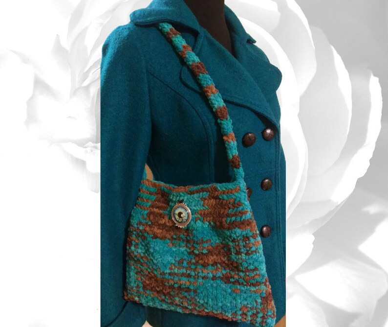 May include: A teal blue and brown crocheted handbag with a decorative button. The bag has a long strap and is worn over the shoulder.