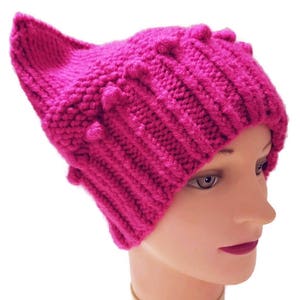 May include: A bright pink knitted hat with a cat ear design. The hat features a ribbed knit pattern and small, textured bobbles along the top. The hat is displayed on a mannequin head.