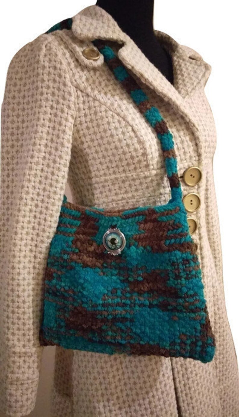 May include: A teal and brown crocheted handbag with a decorative button. The bag is worn over the shoulder of a person wearing a white tweed jacket.