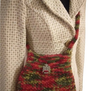 Puede incluir: Un bolso de mano de crochet rojo, verde y naranja con un dije de p&aacute;jaro plateado. El bolso se lleva sobre el hombro de una persona que lleva una chaqueta blanca.