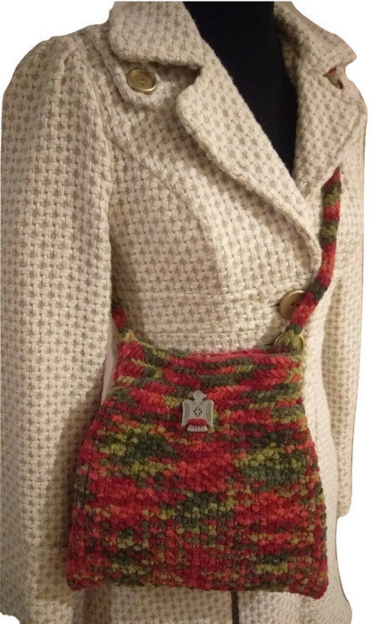 Puede incluir: Un bolso de mano de crochet rojo, verde y naranja con un dije de p&aacute;jaro plateado. El bolso se lleva sobre el hombro de una persona que lleva una chaqueta blanca.