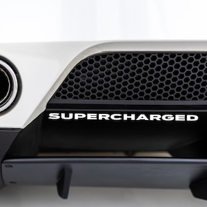 May include: A white car with a black rear bumper and a single chrome exhaust pipe. The bumper has a black honeycomb grille and a white "SUPERCHARGED" decal.