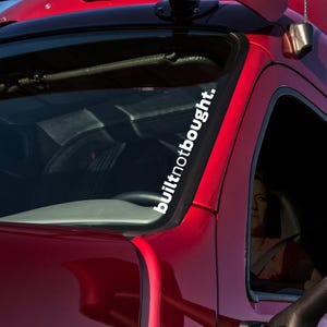 May include: A white vinyl decal with the text "built not bought." The decal is applied to the windscreen of a red lorry.
