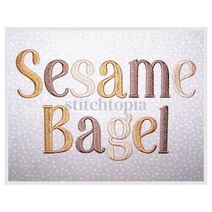 May include: Embroidered text on a white background with a subtle polka dot pattern. The words "Sesame Bagel" are displayed in a mix of gold, brown, and beige thread. The text is in a decorative, stylized font.