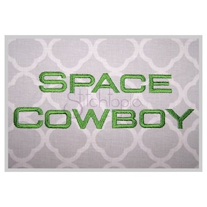 May include: Green embroidered text on a white fabric background that reads "SPACE COWBOY".