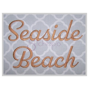 May include: Embroidered design on a light grey fabric background. The design reads "Seaside Beach" in a cursive font with a gold colour.