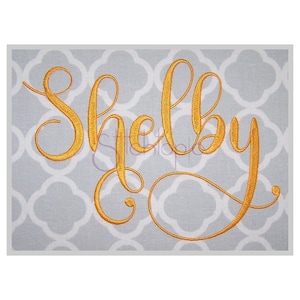 May include: Gold embroidered cursive lettering spelling the name "Shelby" on a gray background.