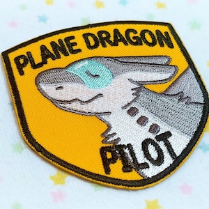 Könnte beinhalten: Ein gelber und schwarzer gestickter Aufnäher mit einem Cartoon-Grauen Drachen mit einem blauen Auge. Der Aufnäher trägt den Text "Plane Dragon Pilot" in schwarzer Schrift.