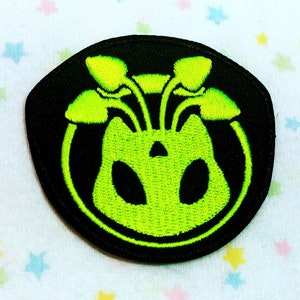 Op de afbeelding: Een geborduurde zwarte en groene patch met een cirkelvormig ontwerp. Het midden van de patch toont een groene, buitenaardse wezens met drie paddenstoelachtige uitgroeisels op zijn hoofd.