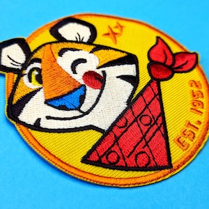 以下が含まれることがあります： 赤いパーティーハットをかぶった漫画のトラが描かれた黄色の刺繍パッチ。トラはウィンクして笑っています。パッチには「EST. 1952」の文字があります。