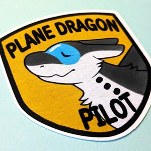 Puede incluir: Una pegatina en forma de escudo amarilla y negra con un dragón de dibujos animados que lleva un casco azul. El texto "PLANE DRAGON PILOT" está escrito en la pegatina.