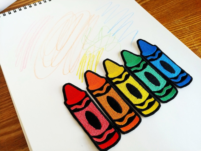 Op de afbeelding: Zes kleurrijke waskrijt-patches in een regenboogvolgorde, beginnend met rood, oranje, geel, groen, blauw en eindigend met paars. De patches bevinden zich op een wit oppervlak.