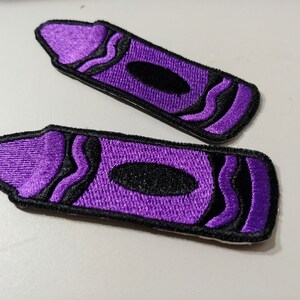 Op de afbeelding: Twee paarse vilt strijk-op patches in de vorm van kleurpotloden. De kleurpotloden hebben zwarte contouren en golvende lijnen.