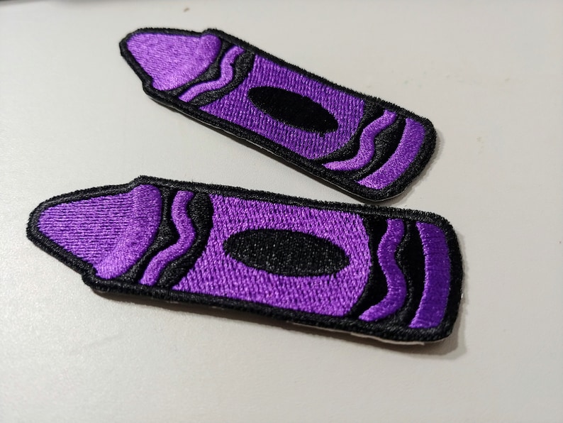 Op de afbeelding: Twee paarse vilt strijk-op patches in de vorm van kleurpotloden. De kleurpotloden hebben zwarte contouren en golvende lijnen.