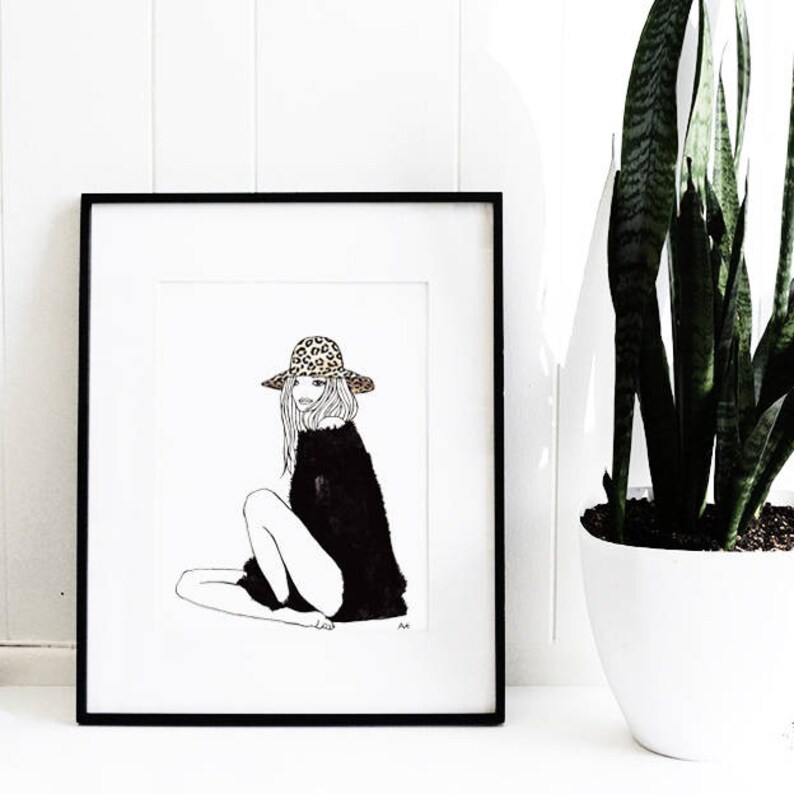May include: Black and white line drawing of a woman wearing a leopard print hat and a black sweater. The drawing is framed in a black frame and is hanging on a white wall.