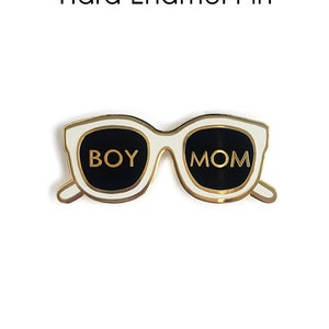 Peut inclure: Épinglette en émail blanc en forme de lunettes de soleil avec des verres noirs. Les verres portent les mots "BOY" et "MOM" écrits en or.