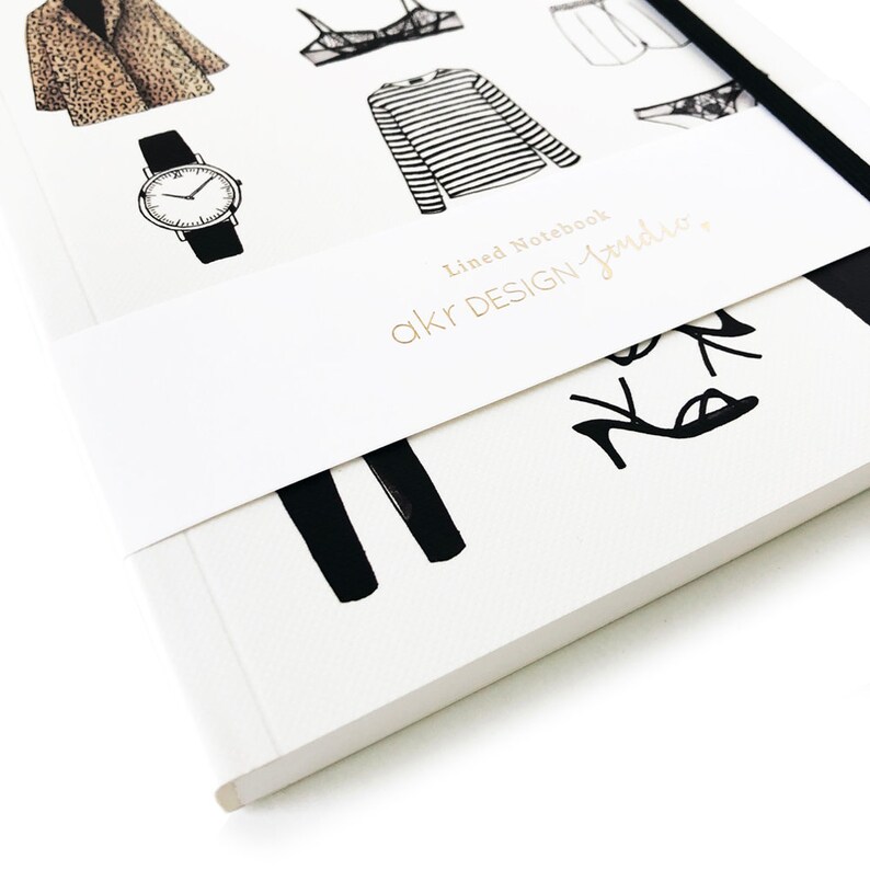 May include: A white notebook with a black and white illustration of clothing and accessories. The notebook has a white band around it with the text "akr DESIGN-studio" in gold lettering. The text "Lined Notebook" is also visible on the notebook.