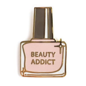 May include: Gold enamel pin shaped like a bottle of nail polish with a pink lid and a white label that says "BEAUTY ADDICT".
