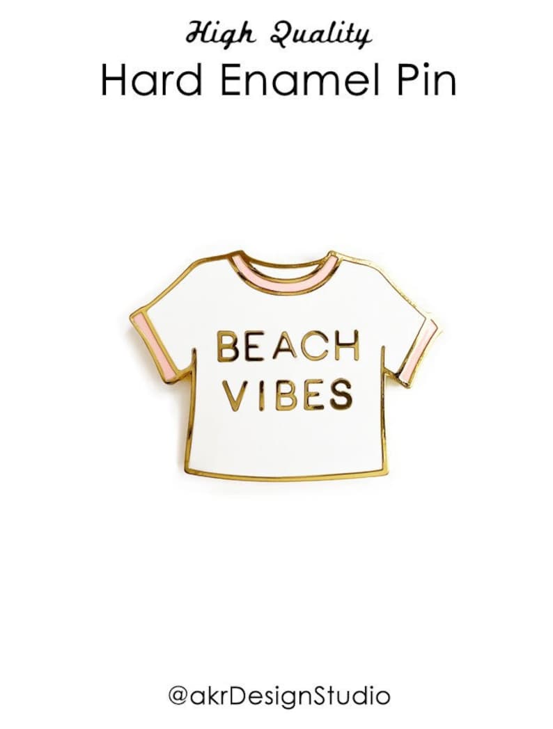 May include: Gold enamel pin shaped like a white t-shirt with pink trim. The t-shirt has the text "BEACH VIBES" printed on it.
