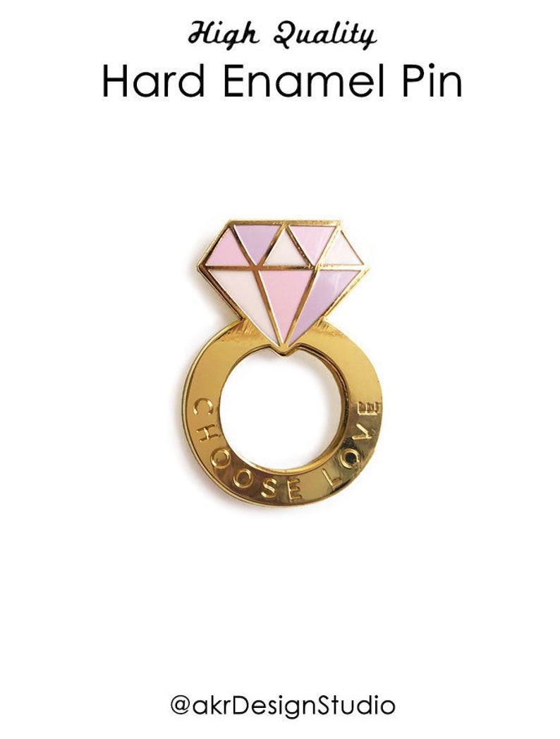 Pu&ograve; includere: Spilla in smalto dorato a forma di anello con un diamante rosa e bianco in cima. L'anello ha le parole "Choose Love" incise.