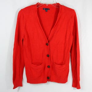 May include: A bright red long-sleeve cardigan featuring a V-neckline, a front button closure with four dark buttons, and two patch pockets at the lower front. The garment includes a Gap brand label at the inner neckline and ribbed cuffs.