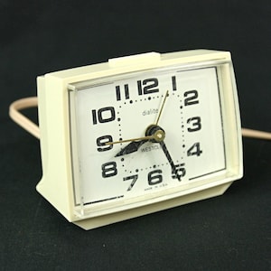 May include: A vintage cream-colored Westclox Dialite alarm clock. The clock has a rectangular face with large black numerals and hands. The clock face is protected by a clear plastic cover. The clock is plugged in and has a power cord.