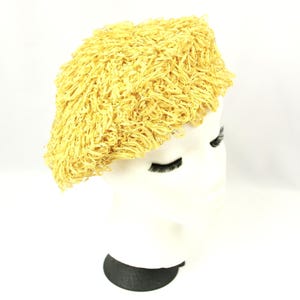 May include: A yellow, textured beret-style hat. The hat is made of a woven material with a fringe-like texture. The hat is displayed on a white mannequin head with black eyelashes and a black base.
