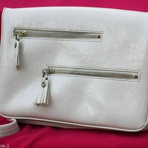 May include: A white leather shoulder bag with two zipped compartments and a flap closure. The bag has a long strap and a small tassel on each zipper pull.