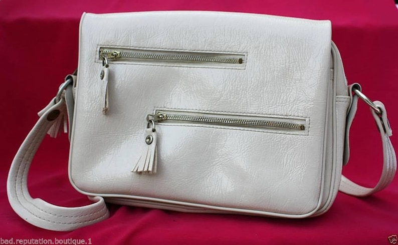 May include: A white leather shoulder bag with two zipped compartments and a flap closure. The bag has a long strap and a small tassel on each zipper pull.