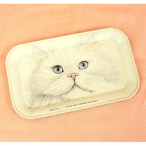 May include: A rectangular, off-white metal tray featuring a detailed illustration of a white cat's face with blue eyes and a pink nose. The tray has rounded corners and a slightly raised edge. Text at the bottom reads "© 1984 MARLEX INTERNATIONAL DESIGN MERLIN".