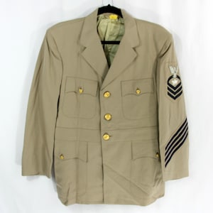 Puede incluir: Chaqueta militar color beige con botones y bolsillos dorados. Cuenta con un emblema en blanco y negro y rayas en la manga. La chaqueta está colgada en una percha negra.