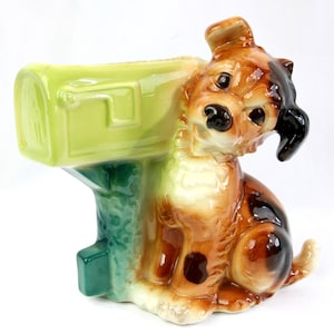 May include: A ceramic dog figurine with a mailbox. The dog is brown, tan, and black with a glossy finish. The mailbox is light green and attached to a green base. The figurine is a decorative item.