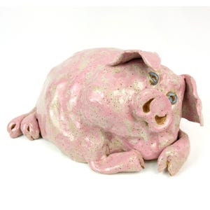 May include: A ceramic pig sculpture in a reclining pose. The pig is primarily pink with speckled, light-colored accents. It has blue eyes and a detailed snout and ears. The sculpture is on a white background.