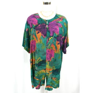 May include: A short-sleeved top with a button-down front, featuring a bold, abstract floral print in green, purple, and orange. The garment is displayed on a mannequin, highlighting its relaxed fit and distinctive design. A gold necklace is visible.