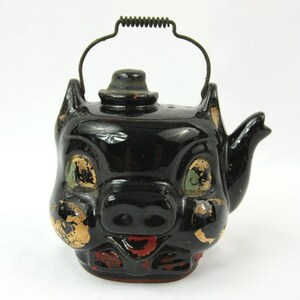 May include: A black ceramic teapot shaped like a pig's head, featuring a handle and spout. The pig has green eyes, gold accents, and a red mouth. The teapot has a small hat on top.