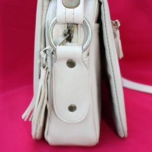 May include: A white leather shoulder bag with a zip closure, a ring detail, and a tassel. The bag has a flap with a snap closure and a small pocket on the front.
